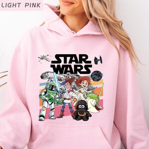 Puede incluir: Sudadera con capucha rosa claro con un gr&aacute;fico de Star Wars con personajes como Boba Fett, Woody y Darth Vader. El logotipo de Star Wars es negro. La sudadera tiene un bolsillo delantero y una capucha con cord&oacute;n.