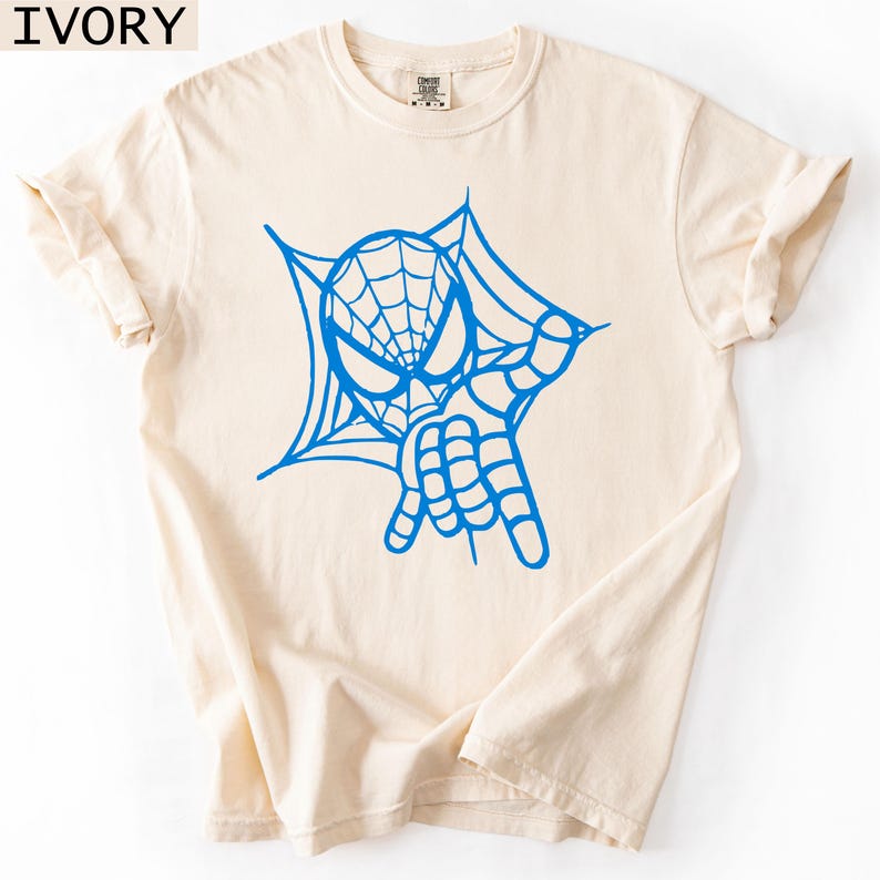 May include: Ivory-colored t-shirt featuring a blue graphic of a superhero's face and hand, with a web design. The shirt has rolled-up sleeves and a crew neck. The word "IVORY" is printed in the top left corner.
