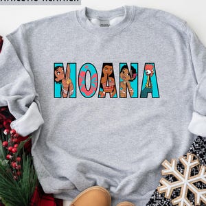 May include: A grey sweatshirt featuring the word "MOANA" in a colourful graphic design. The letters are filled with images of characters from the animated film. The text "ATHLETIC HEATHER" is printed at the top.