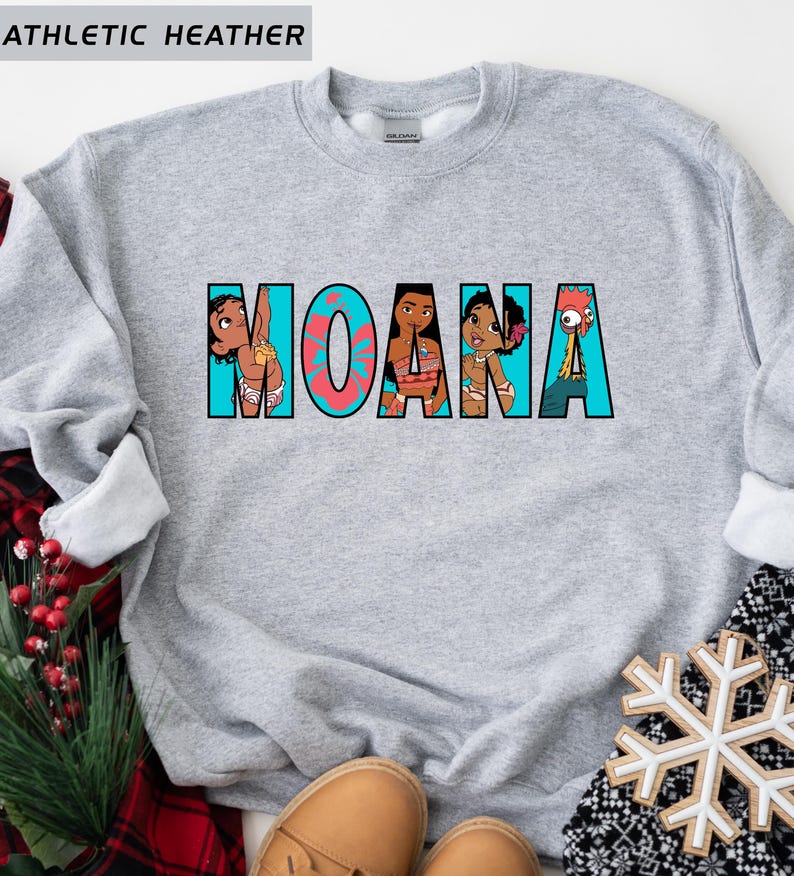 May include: A grey sweatshirt featuring the word "MOANA" in a colourful graphic design. The letters are filled with images of characters from the animated film. The text "ATHLETIC HEATHER" is printed at the top.
