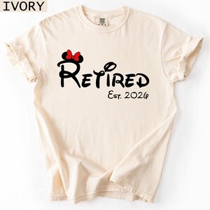 Puede incluir: Camiseta de color marfil con la palabra "Retired" en una fuente caprichosa, acompa&ntilde;ada de "Est. 2026". Una silueta negra de la cabeza de Minnie Mouse con un lazo rojo est&aacute; encima de la palabra "Retired".