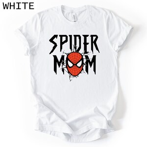 May include: White t-shirt featuring the text "SPIDER MOM" in black, with a red and black spider-themed graphic. The graphic includes a spider mask design. The word "WHITE" is in the top left corner.