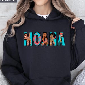 May include: Black hooded sweatshirt featuring the word "MOANA" in turquoise and coral lettering. The letters are filled with cartoon illustrations of characters from the film. The word "BLACK" is in a white box in the upper left corner.