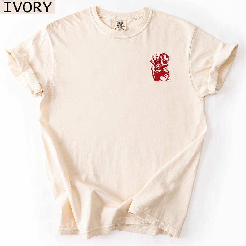 Puede incluir: Una camiseta de color marfil de manga corta y cuello redondo con un peque&ntilde;o gr&aacute;fico rojo de un personaje rob&oacute;tico en la parte superior izquierda del pecho. La prenda tiene un corte est&aacute;ndar con cuello acanalado y el texto IVORY es visible.