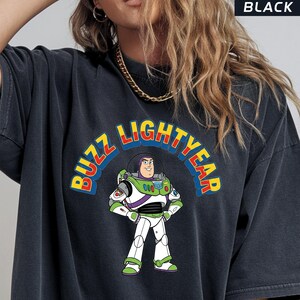 Op de afbeelding: Zwart T-shirt met een afbeelding van Buzz Lightyear uit Toy Story. Het ontwerp bevat de woorden "BUZZ LIGHTYEAR" in een kleurrijke boog boven het personage. Het shirt heeft een donkergrijze kleur.