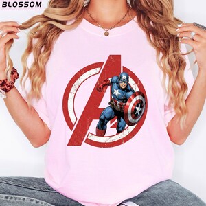 Puede incluir: Camiseta rosa claro con el logotipo de los Vengadores en rojo desgastado y el Capit&aacute;n Am&eacute;rica. El gr&aacute;fico muestra al Capit&aacute;n Am&eacute;rica con su escudo. La palabra "BLOSSOM" est&aacute; en la parte superior izquierda.
