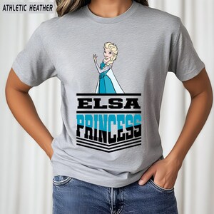 May include: Athletic heather gray t-shirt featuring a graphic of Elsa from Frozen. The design includes the text "ELSA PRINCESS" in black and blue. The image of Elsa is in a blue dress, waving.