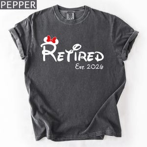 Puede incluir: Una camiseta gris oscuro con la palabra "Retired" en escritura blanca, con un lazo de Minnie Mouse y el texto "Est. 2026". La camiseta tiene mangas enrolladas y cuello redondo.