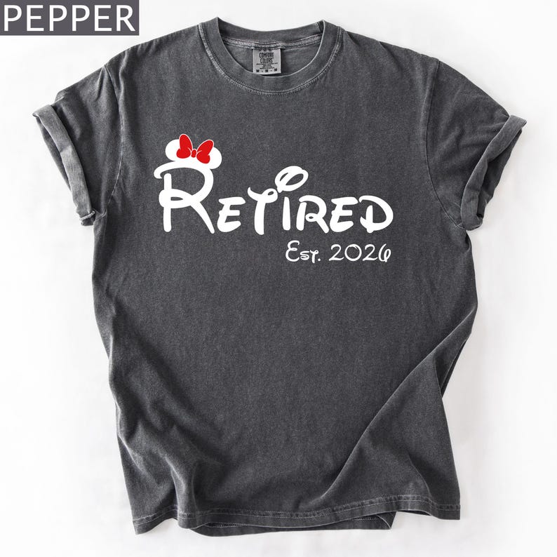 Puede incluir: Una camiseta gris oscuro con la palabra "Retired" en escritura blanca, con un lazo de Minnie Mouse y el texto "Est. 2026". La camiseta tiene mangas enrolladas y cuello redondo.