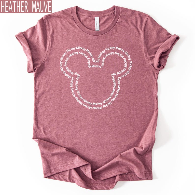 Peut inclure: T-shirt mauve chin&eacute; avec un motif de t&ecirc;te de Mickey Mouse blanc. La t&ecirc;te de Mickey Mouse est form&eacute;e par le mot "Mickey" r&eacute;p&eacute;t&eacute; en cercle. Le t-shirt est fait d'une mati&egrave;re douce.