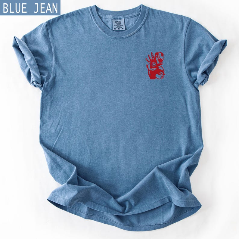 Puede incluir: Una camiseta de manga corta color azul jean con un peque&ntilde;o gr&aacute;fico rojo de un personaje de superh&eacute;roe en el &aacute;rea del pecho izquierdo. La prenda tiene cuello redondo y una etiqueta en el cuello interior. El texto BLUE JEAN es visible.