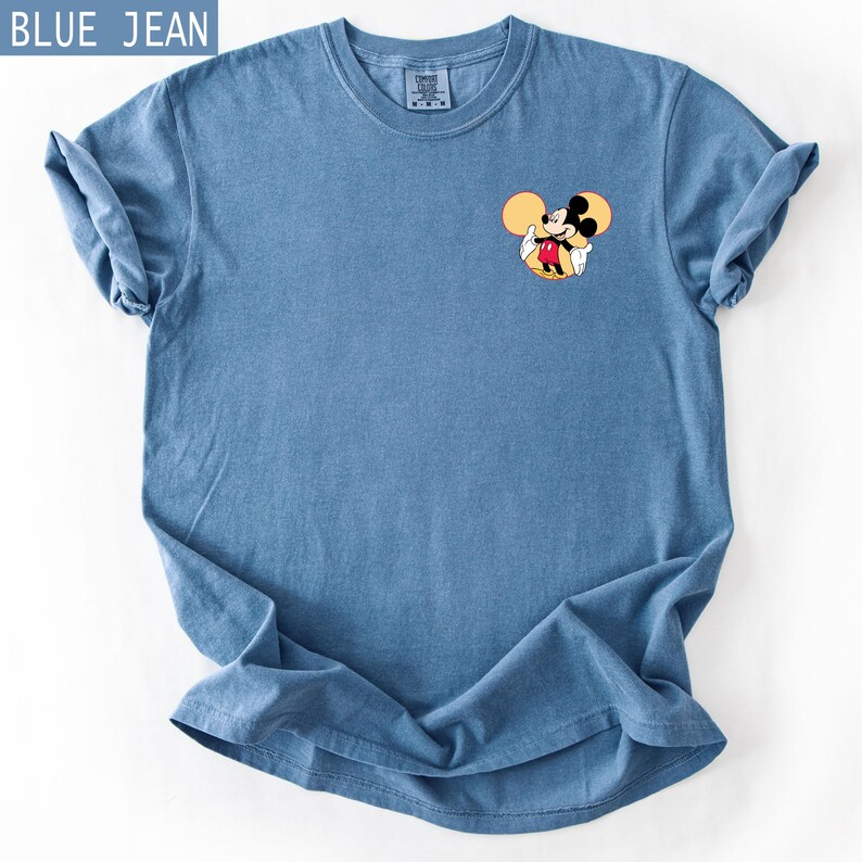 May include: Blue jean-coloured t-shirt with a small Mickey Mouse graphic on the front. The graphic shows Mickey Mouse in his classic outfit. The shirt has a crew neck and short sleeves. The text "BLUE JEAN" is in the upper left corner.