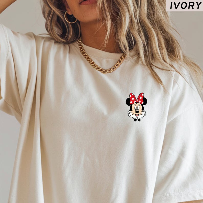 May include: Ivory-coloured t-shirt with a Minnie Mouse graphic. The graphic shows Minnie Mouse with a red polka-dot bow. The shirt has short sleeves and a crew neck. The word "IVORY" is printed at the top.