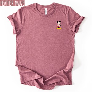 May include: A heather mauve short-sleeved t-shirt featuring a small graphic of Mickey Mouse on the upper left chest. The character is depicted in his classic red shorts, white gloves, and yellow shoes. The shirt has a crew neckline and a soft texture.