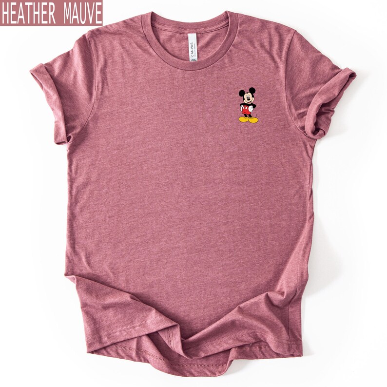 May include: A heather mauve short-sleeved t-shirt featuring a small graphic of Mickey Mouse on the upper left chest. The character is depicted in his classic red shorts, white gloves, and yellow shoes. The shirt has a crew neckline and a soft texture.