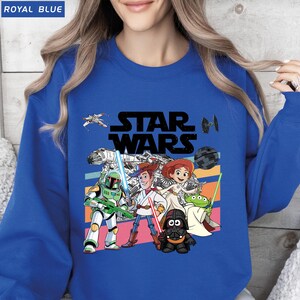 Puede incluir: Sudadera azul real con un gr&aacute;fico de Star Wars que presenta personajes de la franquicia. El dise&ntilde;o incluye a Woody, Buzz Lightyear y Darth Vader, con un fondo de rayas retro. El texto "ROYAL BLUE" est&aacute; en la esquina superior izquierda.