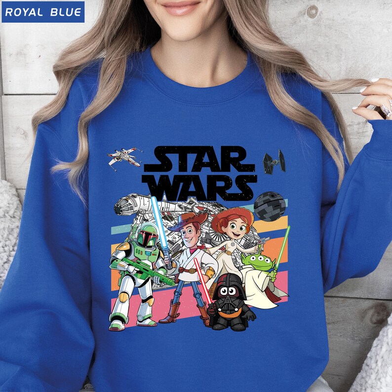 Puede incluir: Sudadera azul real con un gr&aacute;fico de Star Wars que presenta personajes de la franquicia. El dise&ntilde;o incluye a Woody, Buzz Lightyear y Darth Vader, con un fondo de rayas retro. El texto "ROYAL BLUE" est&aacute; en la esquina superior izquierda.