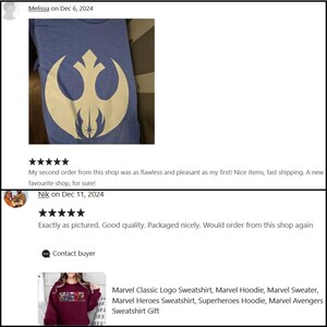 May include: A blue t-shirt with a cream-coloured graphic design. The design is a stylised emblem with a circular shape and a central starburst. The shirt is folded, with the design prominently displayed.