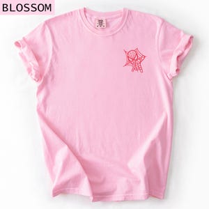 K&ouml;nnte beinhalten: Hellrosa T-Shirt mit hochgekrempelten &Auml;rmeln. Auf der Vorderseite ist eine rote Spider-Man-Grafik aufgedruckt. Das Wort "BLOSSOM" befindet sich oben links. Das Shirt besteht aus einem weichen Material.
