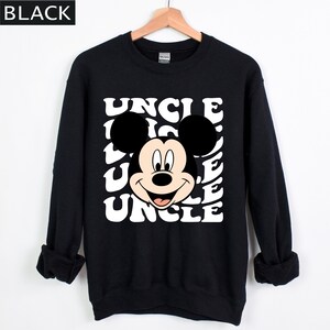 Pu&ograve; includere: Felpa nera con il volto sorridente di Topolino e la parola "UNCLE" in un motivo ripetuto. L'indumento &egrave; esposto su una gruccia di legno su sfondo bianco. La parola "BLACK" si trova nell'angolo in alto a sinistra.