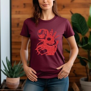 Puede incluir: Camiseta granate con un dise&ntilde;o gr&aacute;fico de purpurina roja. El gr&aacute;fico parece ser una criatura o s&iacute;mbolo estilizado. La camiseta es de color liso y tiene cuello redondo.
