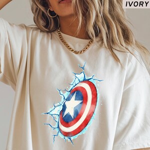 May include: Ivory-colored t-shirt featuring a graphic of Captain America's shield, with red, white, and blue colors. The shield design appears to be breaking through the shirt, with blue crack lines. The word "IVORY" is in the top right corner.