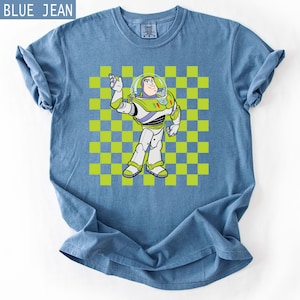 May include: Blue jean t-shirt featuring a cartoon image of Buzz Lightyear from Toy Story. The character is depicted in his signature space ranger uniform, standing in front of a green and yellow checkered background. The shirt has rolled-up sleeves.