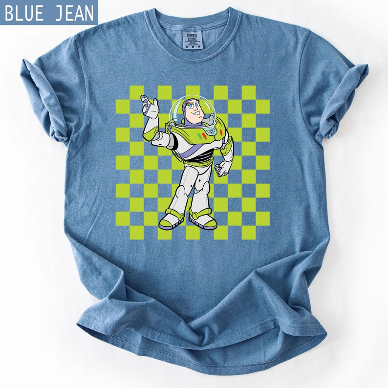 May include: Blue jean t-shirt featuring a cartoon image of Buzz Lightyear from Toy Story. The character is depicted in his signature space ranger uniform, standing in front of a green and yellow checkered background. The shirt has rolled-up sleeves.