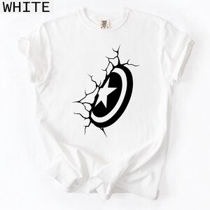 Pode incluir: Camiseta branca com um gr&aacute;fico preto e branco de um escudo com uma estrela, parecendo romper a superf&iacute;cie. A palavra "WHITE" est&aacute; impressa no canto superior esquerdo. Gola redonda, mangas curtas. Roupa casual.