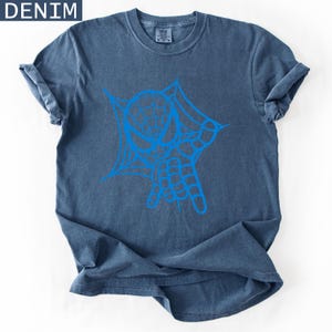 May include: Denim blue t-shirt with a blue graphic of a spider web and hand. The t-shirt has rolled-up sleeves and a crew neck. The word "DENIM" is in the upper left corner.