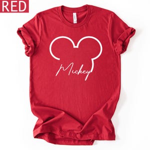 May include: A red t-shirt with a white outline of Mickey Mouse's head and the name "Mickey" in a cursive font. The shirt has rolled-up sleeves and a crew neck. The word "RED" is in a red box in the upper left corner.
