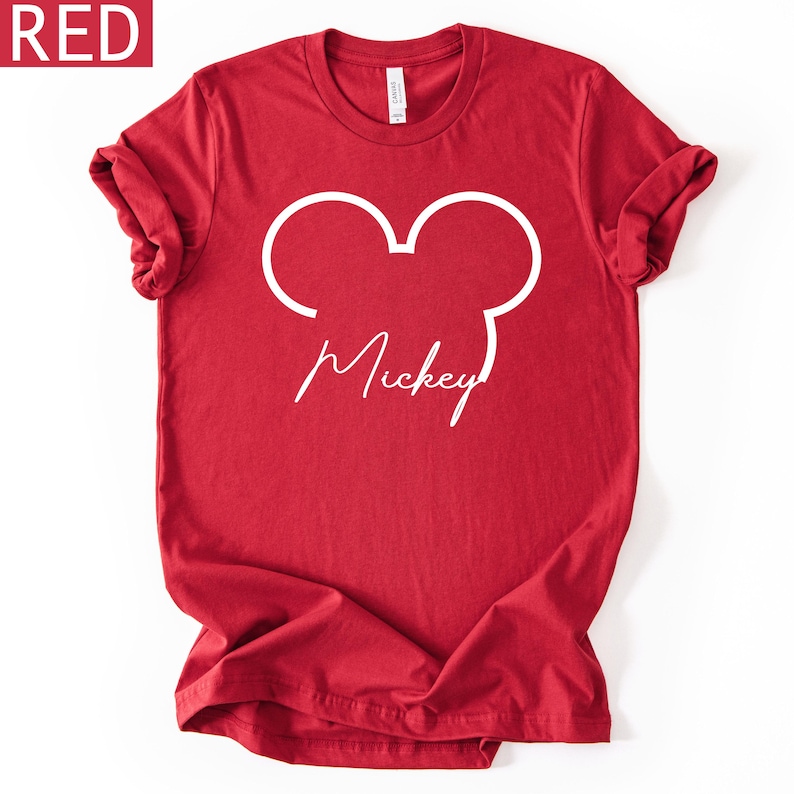 May include: A red t-shirt with a white outline of Mickey Mouse's head and the name "Mickey" in a cursive font. The shirt has rolled-up sleeves and a crew neck. The word "RED" is in a red box in the upper left corner.