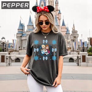 May include: Dark gray t-shirt featuring a graphic of characters from the movie Frozen, surrounded by snowflakes and bows. The word "PEPPER" is in the upper left corner. The person is wearing a Minnie Mouse ear headband and sunglasses.
