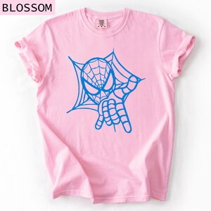 May include: A light pink t-shirt featuring a blue graphic of a spider-webbed mask and hand gesture. The word "BLOSSOM" is printed at the top left. The shirt has rolled-up sleeves.