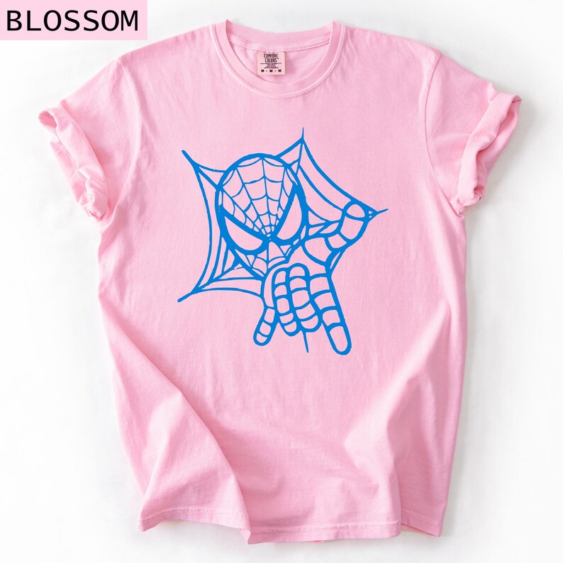 May include: A light pink t-shirt featuring a blue graphic of a spider-webbed mask and hand gesture. The word "BLOSSOM" is printed at the top left. The shirt has rolled-up sleeves.