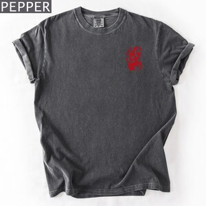 Puede incluir: Una camiseta de manga corta en color gris carb&oacute;n con un acabado desgastado de estilo vintage. La prenda presenta un peque&ntilde;o estampado gr&aacute;fico rojo en la parte superior izquierda del pecho. La palabra PEPPER aparece en blanco arriba.