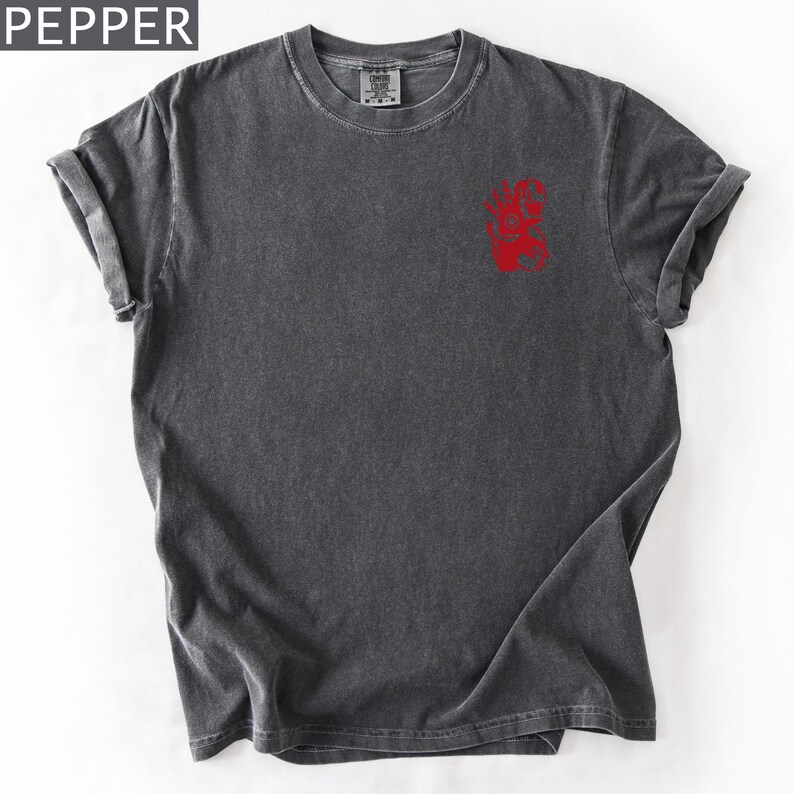 Puede incluir: Una camiseta de manga corta en color gris carb&oacute;n con un acabado desgastado de estilo vintage. La prenda presenta un peque&ntilde;o estampado gr&aacute;fico rojo en la parte superior izquierda del pecho. La palabra PEPPER aparece en blanco arriba.