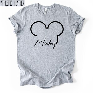 May include: Athletic heather grey t-shirt featuring a black outline of Mickey Mouse ears and the name "Mickey" in a cursive font. The shirt is made of a soft, comfortable material.