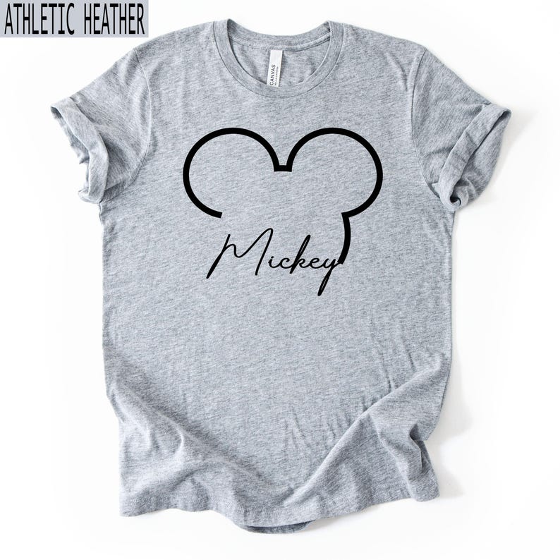 May include: Athletic heather grey t-shirt featuring a black outline of Mickey Mouse ears and the name "Mickey" in a cursive font. The shirt is made of a soft, comfortable material.