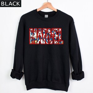 Puede incluir: Sudadera negra con la palabra "MARVEL" en letras rojas con contorno. Las letras est&aacute;n rellenas de im&aacute;genes de Spider-Man en rojo y azul. La sudadera tiene mangas largas y cuello redondo.