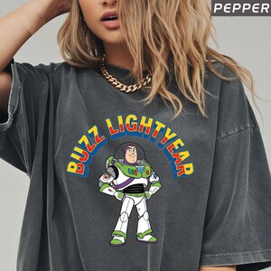 Op de afbeelding: Donkergrijs T-shirt met een kleurrijke afbeelding van Buzz Lightyear en de tekst "BUZZ LIGHTYEAR" in een gebogen ontwerp. Het stripfiguur draagt zijn witte, groene en paarse ruimtepak.