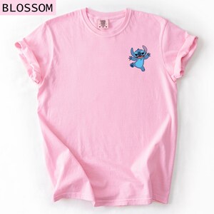 May include: A light pink t-shirt featuring a small blue cartoon character graphic on the front. The word "BLOSSOM" is printed at the top left corner of the image. The shirt has rolled-up sleeves.
