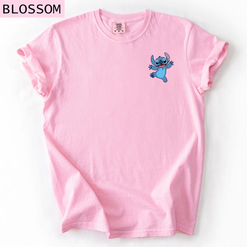 May include: A light pink t-shirt featuring a small blue cartoon character graphic on the front. The word "BLOSSOM" is printed at the top left corner of the image. The shirt has rolled-up sleeves.