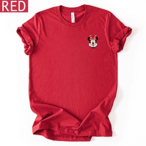 May include: A solid red short-sleeved t-shirt featuring a small graphic of Minnie Mouse on the upper left chest. The character is depicted with her signature red and white polka dot bow, black ears, and white gloves resting under her chin.