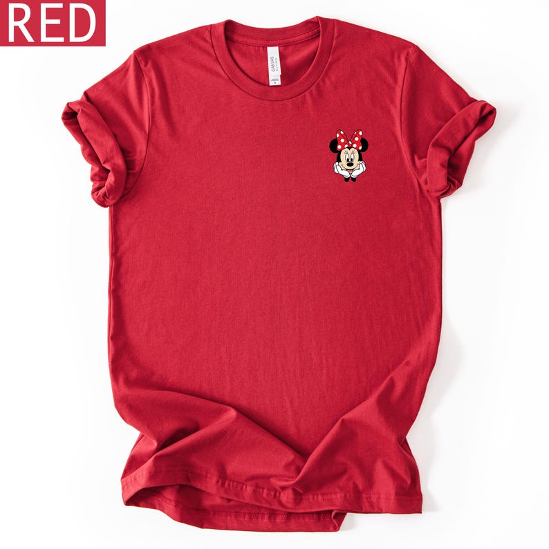 May include: A solid red short-sleeved t-shirt featuring a small graphic of Minnie Mouse on the upper left chest. The character is depicted with her signature red and white polka dot bow, black ears, and white gloves resting under her chin.