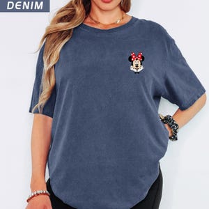 May include: Denim blue t-shirt with a small Minnie Mouse graphic on the chest. The graphic shows Minnie Mouse with a red bow and white gloves. The shirt has short sleeves and a crew neck.