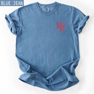 K&ouml;nnte beinhalten: Blaues Jeans-T-Shirt mit roter Grafik auf der Vorderseite. Das Shirt hat einen Rundhalsausschnitt und kurze &Auml;rmel, wobei die &Auml;rmel hochgekrempelt sind. Der Text "BLUE JEAN" befindet sich in einem blauen Feld oben links.