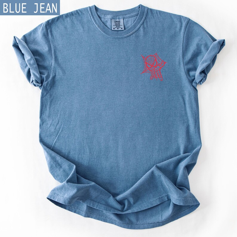 K&ouml;nnte beinhalten: Blaues Jeans-T-Shirt mit roter Grafik auf der Vorderseite. Das Shirt hat einen Rundhalsausschnitt und kurze &Auml;rmel, wobei die &Auml;rmel hochgekrempelt sind. Der Text "BLUE JEAN" befindet sich in einem blauen Feld oben links.