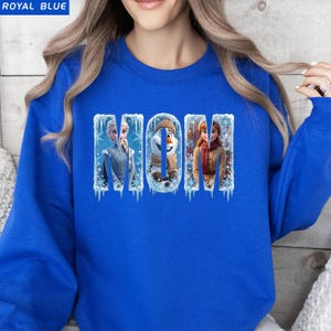 Puede incluir: Sudadera azul real con la palabra "MOM" en una fuente invernal. Las letras est&aacute;n rellenas de im&aacute;genes de personajes de la pel&iacute;cula Frozen, incluyendo Elsa, Olaf y Anna. La sudadera es de color liso.