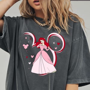 May include: Dark gray t-shirt featuring a cartoon illustration of Ariel from The Little Mermaid in a pink dress. The design includes a Mickey Mouse ear outline with the number 30, stars, and heart-shaped Mickey heads. The word "PEPPER" is at the top.